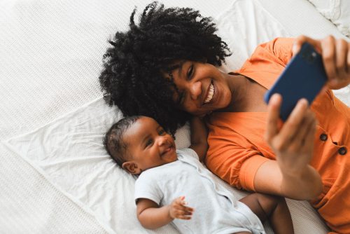 african american woman wearing a orange shirt and her baby lying on a bed taking a picture together
